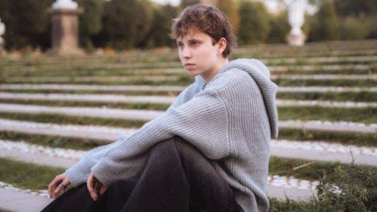 A serious teen in a gray sweater sitting on a staircase outside.