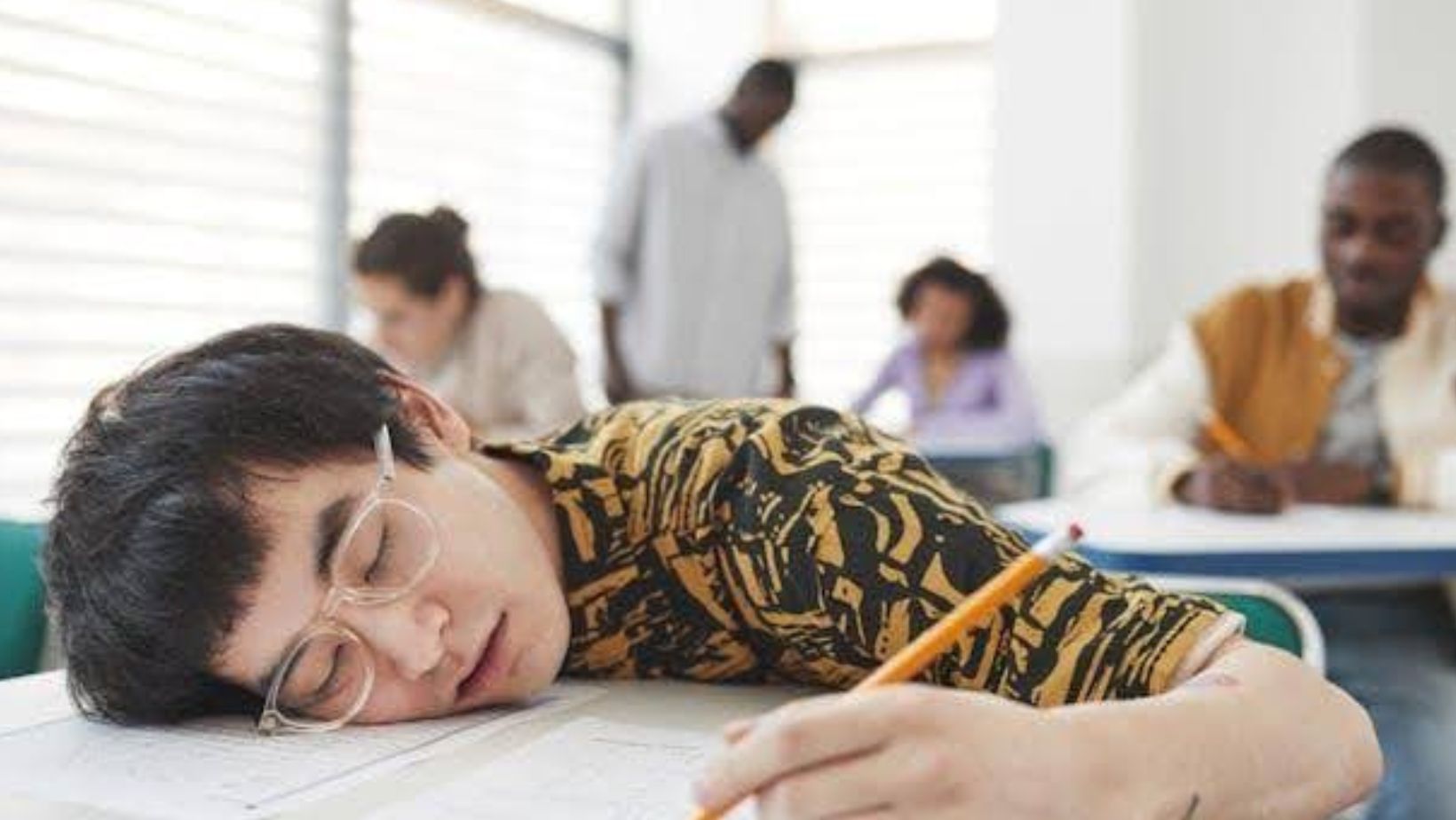 A teen with glasses falling asleep in class at school.